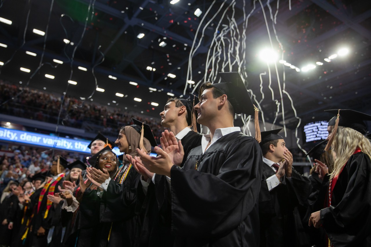 Students smiling and wearing graduation caps and gowns while clapping in arena while confetti falls