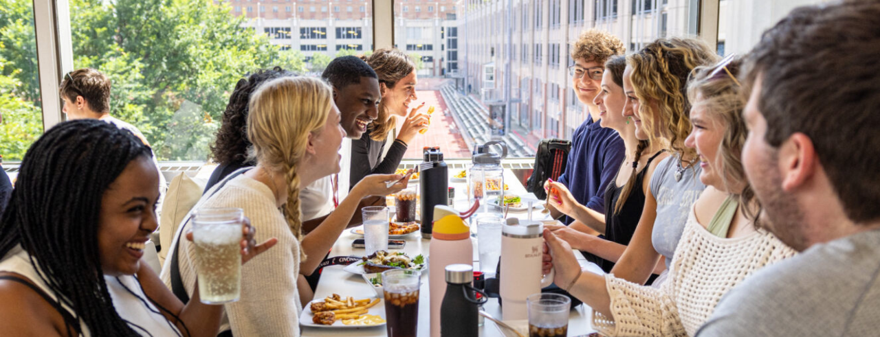 A long table in a dining hall filled with students eating. The students are smiling as they appear to be chatting with each other.