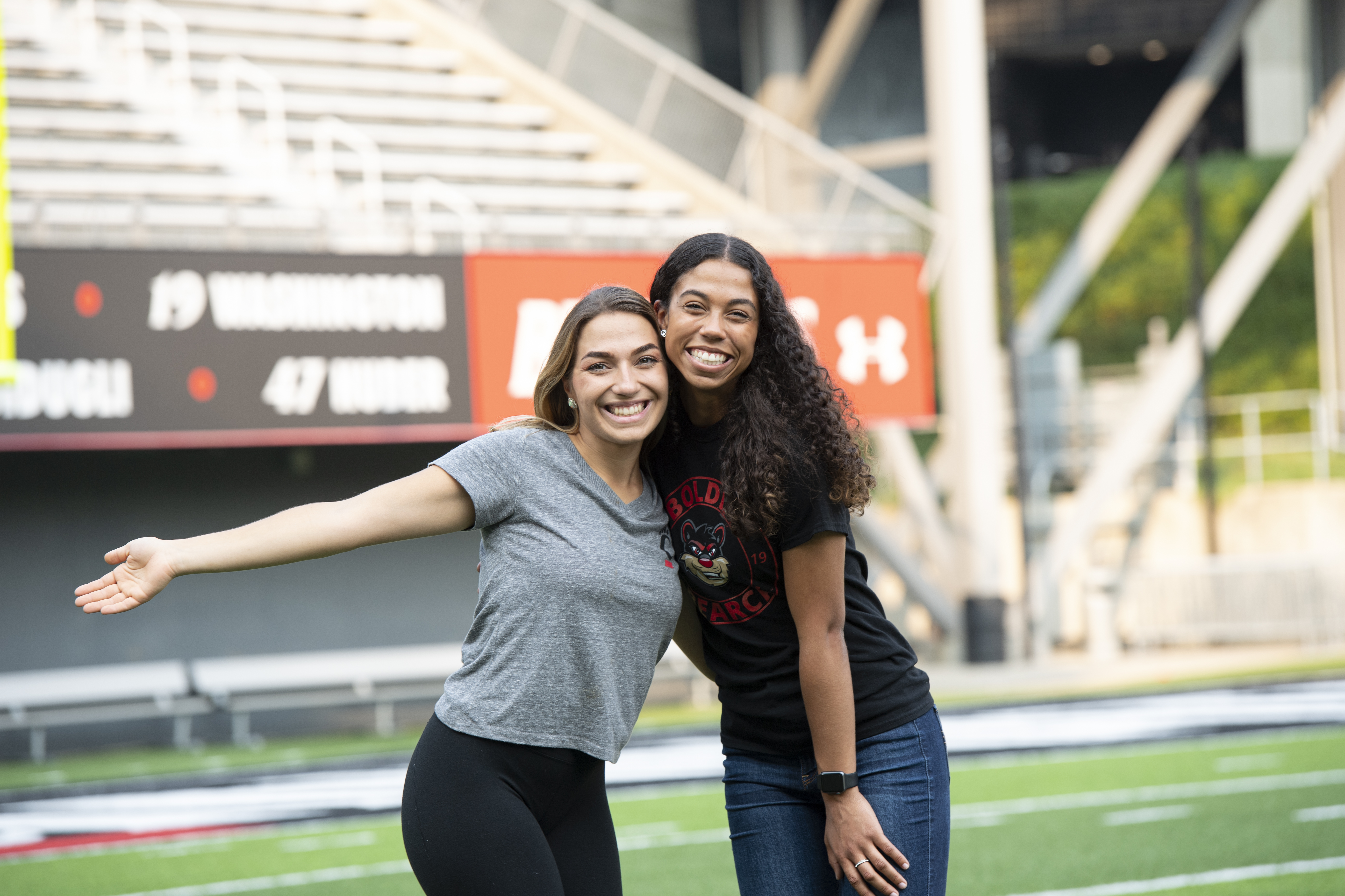 two female UC students standing on the field at Nippert Stadium with seating in the background.