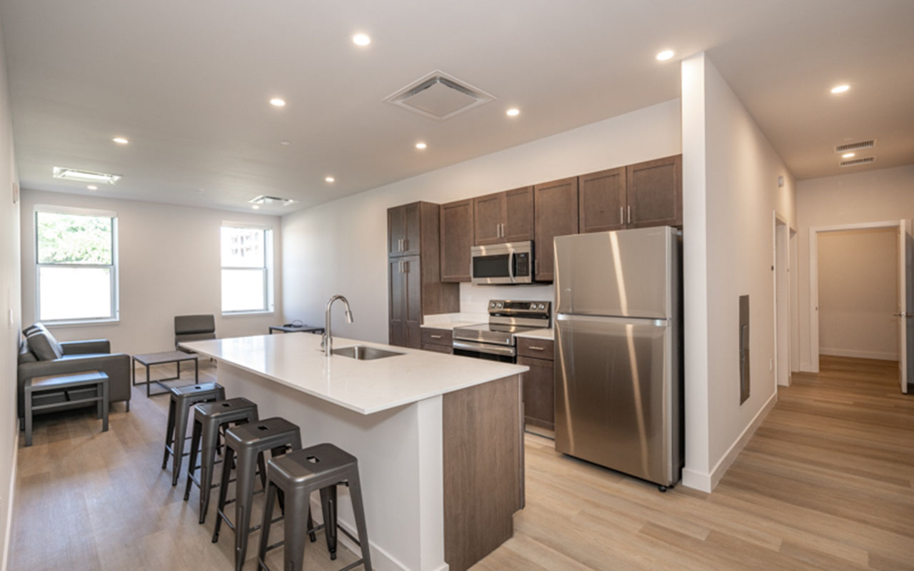 Shared kitchen with refrigerator, cooktop and high counters with chairs, living area in background