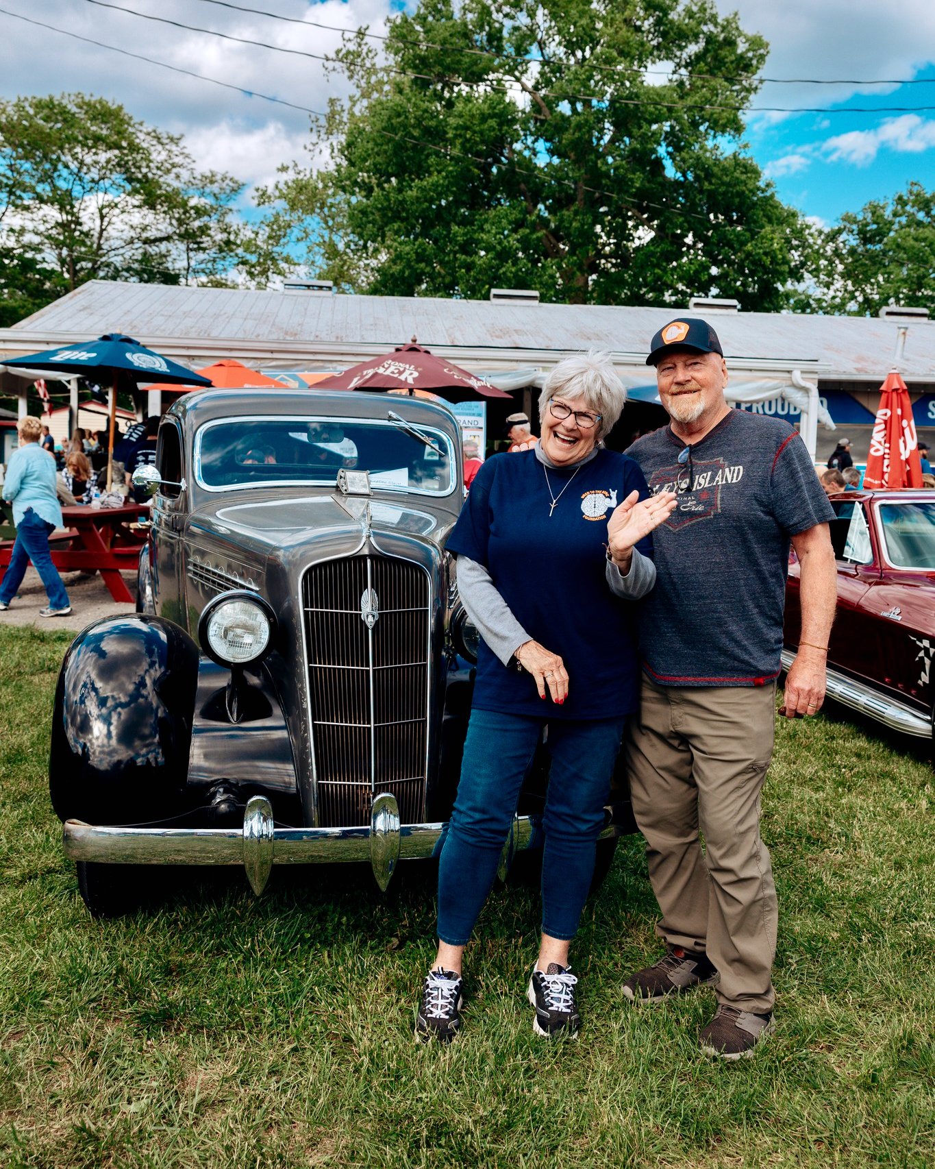 A woman and man stand in front of a vintage car at a classic car show.