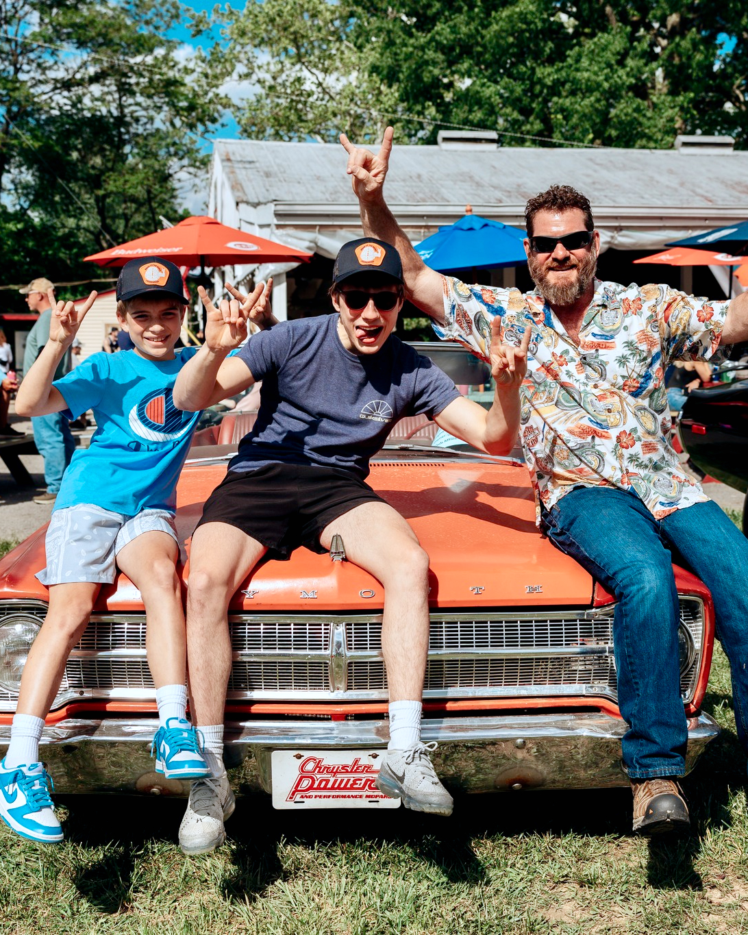 Two boys and a man sit on the hood of an orange classic car in a grassy area of a car show.