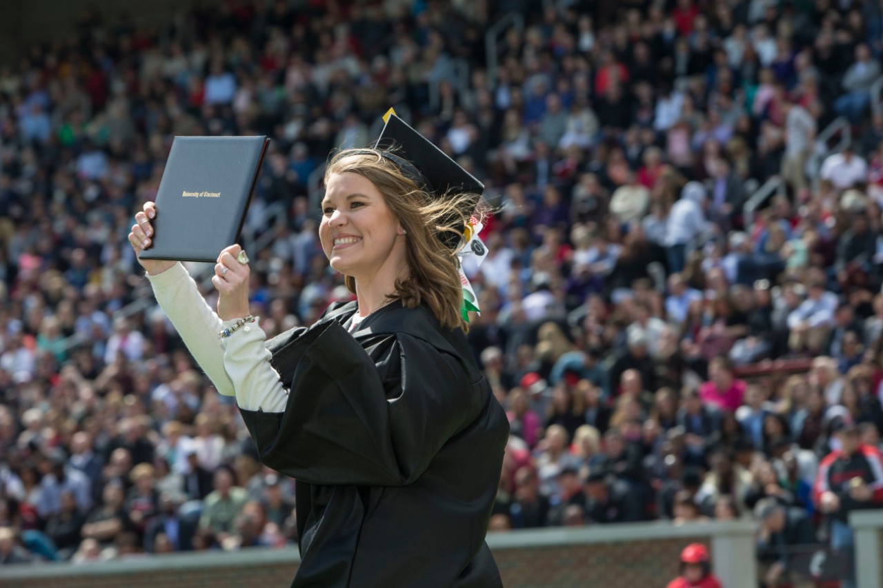 A UC grad in a black cap and gown raises holds up her diploma for supporters to see as she walks off the stage.