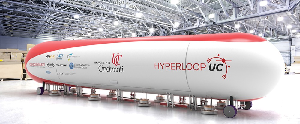 a white and red tubular pod labeled "University of CIncinnati HyperloopUC" sits inside a brightly lit warehouse space with metal rafters