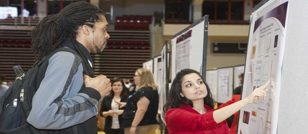 Student presenting a research poster to another student holding a  bookbag with other people in the background.