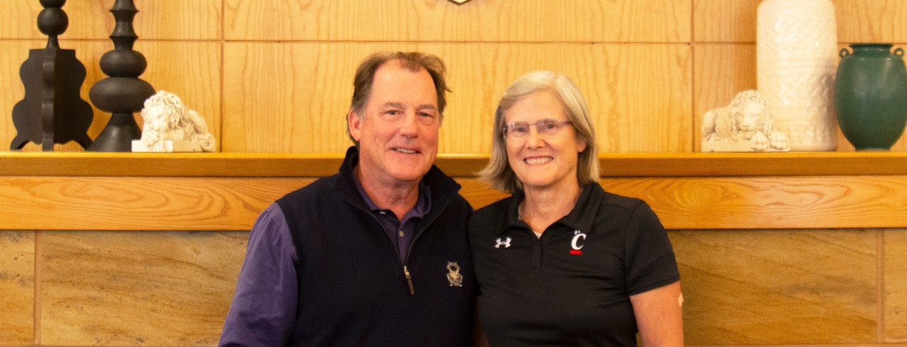 Dick and Cornelia Thornburgh wearing dark clothing with the UC Athletics logo stand and smile in front of a tan wall from a 2019 visit to campus