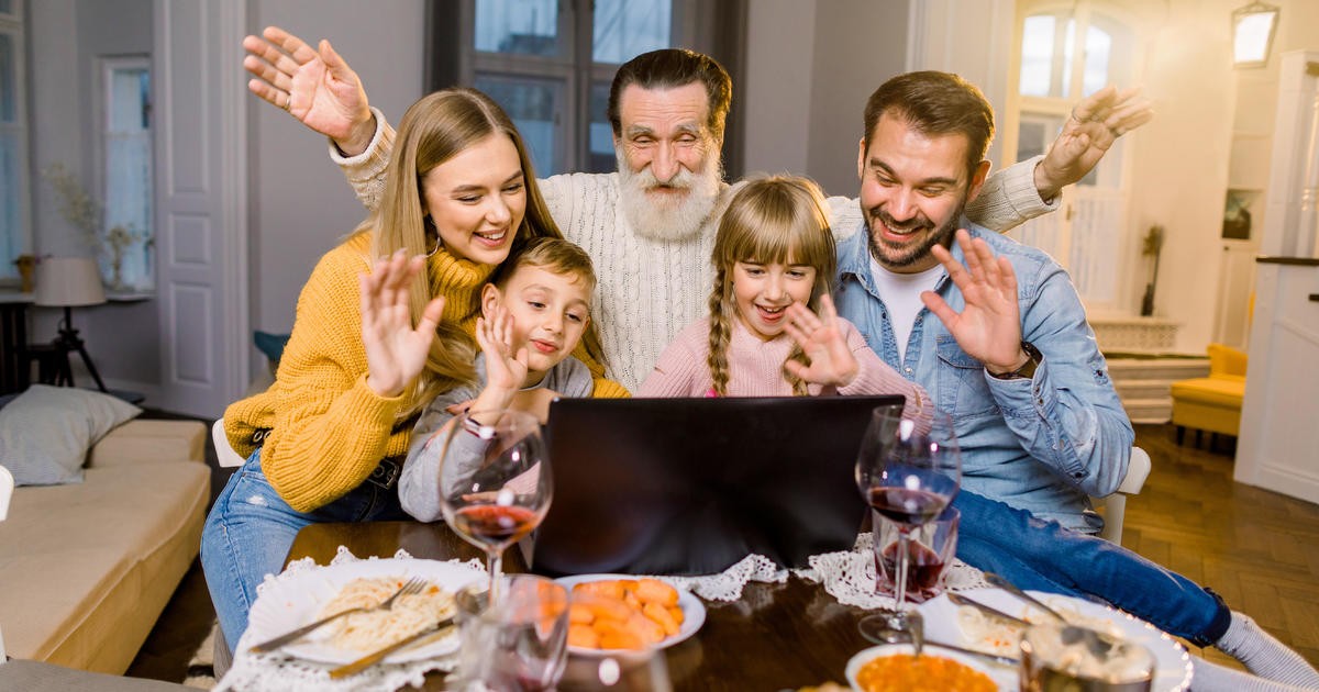A family at a thanksgiving dinner smiling and waving at a laptop on the table