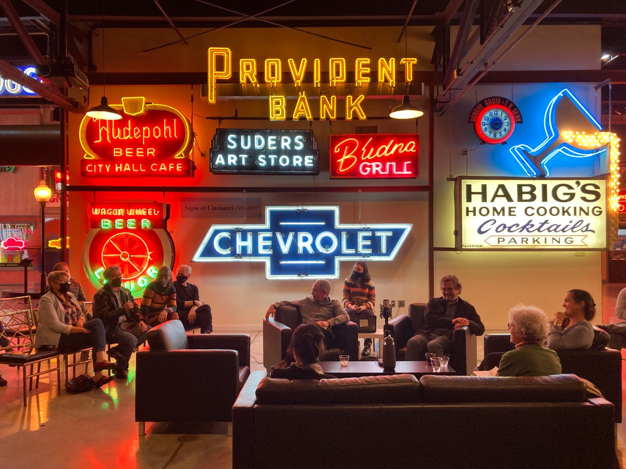 Guest speakers sitting around a coffee table talk in front of neon signs at the American Sign Museum.