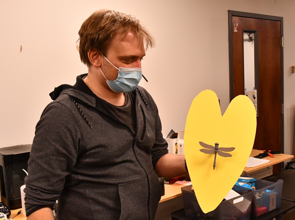 Dieter Vanderelst in a face mask holds up a 3D printed dragonfly mounted to a leaf made from yellow construction paper.