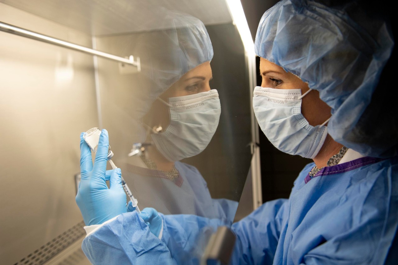 UC College of Pharmacy pharmacist Mary Burns in a mask and gown draws a vial of COVID-19 vaccine under a glass hood with her reflection.