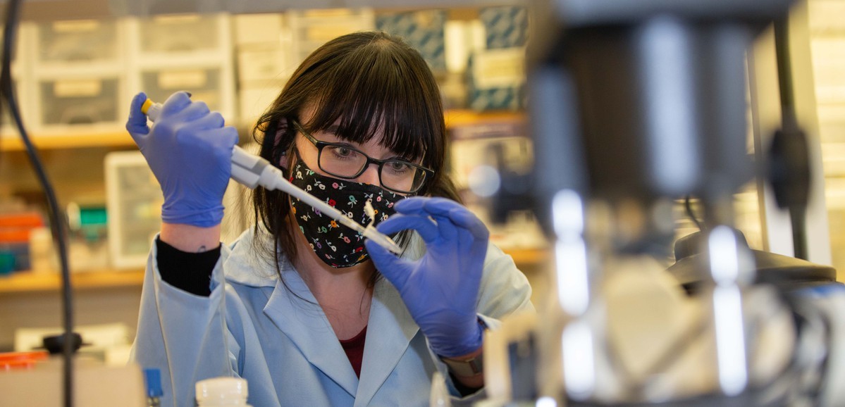 A researcher in gloves and a mask works at a lab bench.