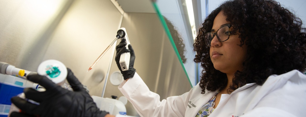 Amber Amparo, wearing a white lab coat and gloves, pipettes samples in the laboratory