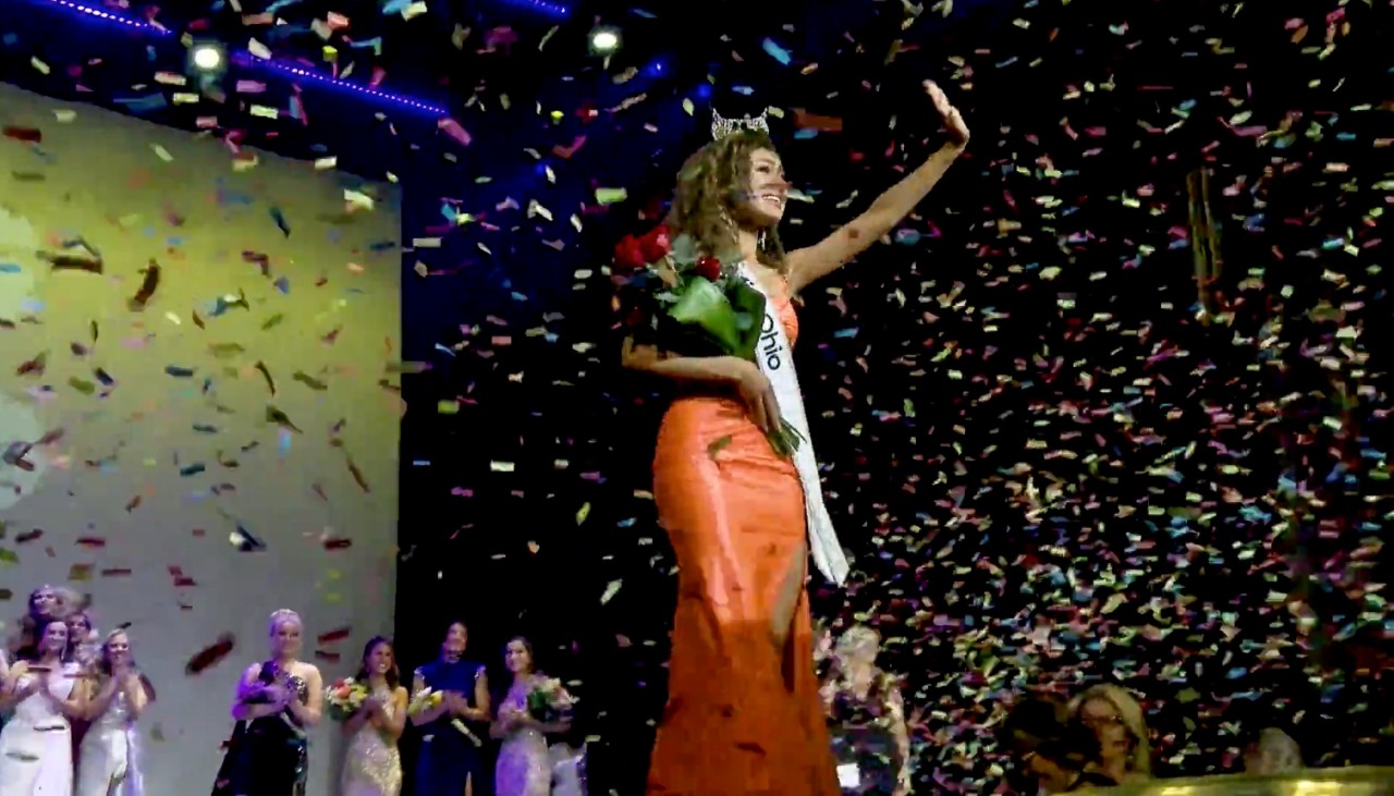 Stephanie Finoti waves to supporters while holding a bouquet of roses as confetti falls on stage.