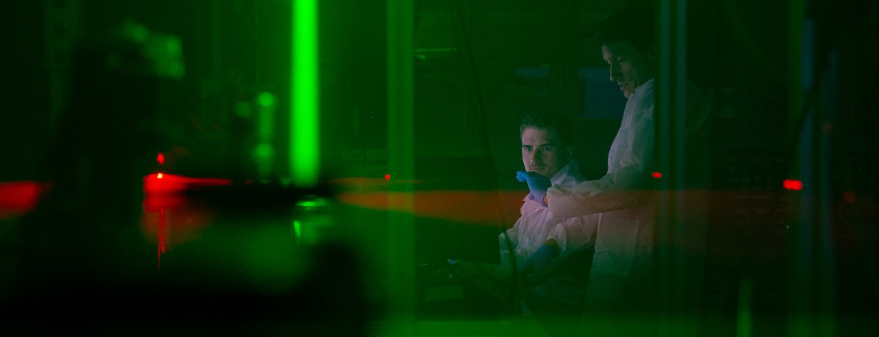 A green laser shines in a dark room while two researchers wearing white lab coats talk in the background