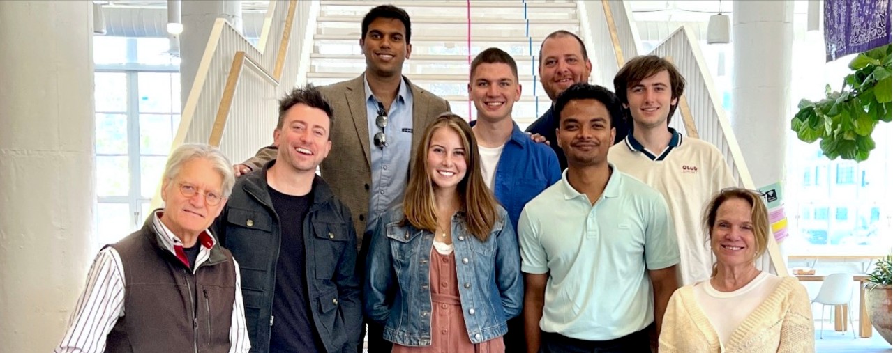 Nine men and women stand together at the bottom of a stairway facing forward smiling.