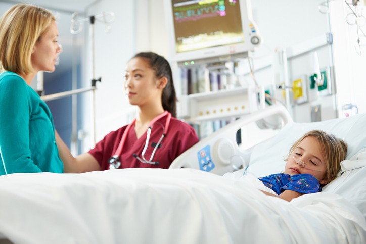 A child sleeps in a hospital bed while a nurse and mother stand and have a conversation