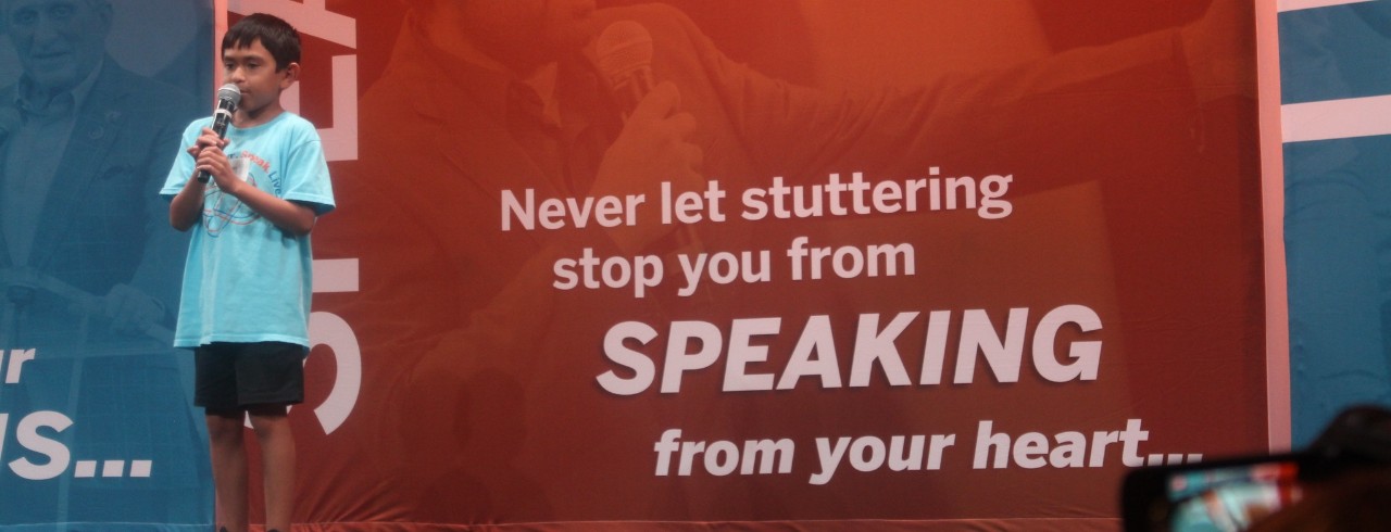 A child on a stage speaks into a microphone with the words “Never let stuttering stop you from SPEAKING from your heart” in the background.