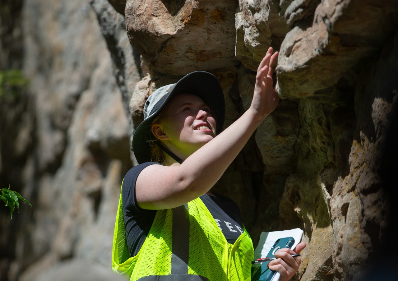 A UC student in a yellow safety vest examines the face of a cliff.