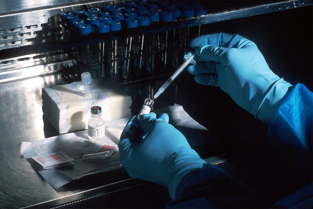 A person’s hands, wearing gloves, pushes a syringe into a vial.