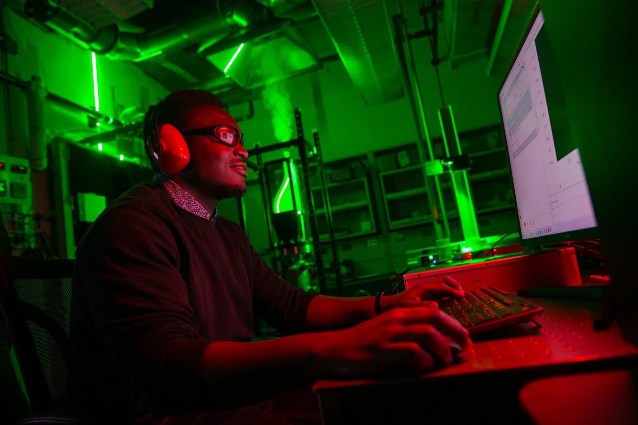 Vincent Onoja sits at a computer while a laser fires at a window in a jet engine behind him.