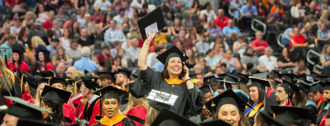 A UC graduate in a cap and gown waves from the floor of Fifth Third Arena.