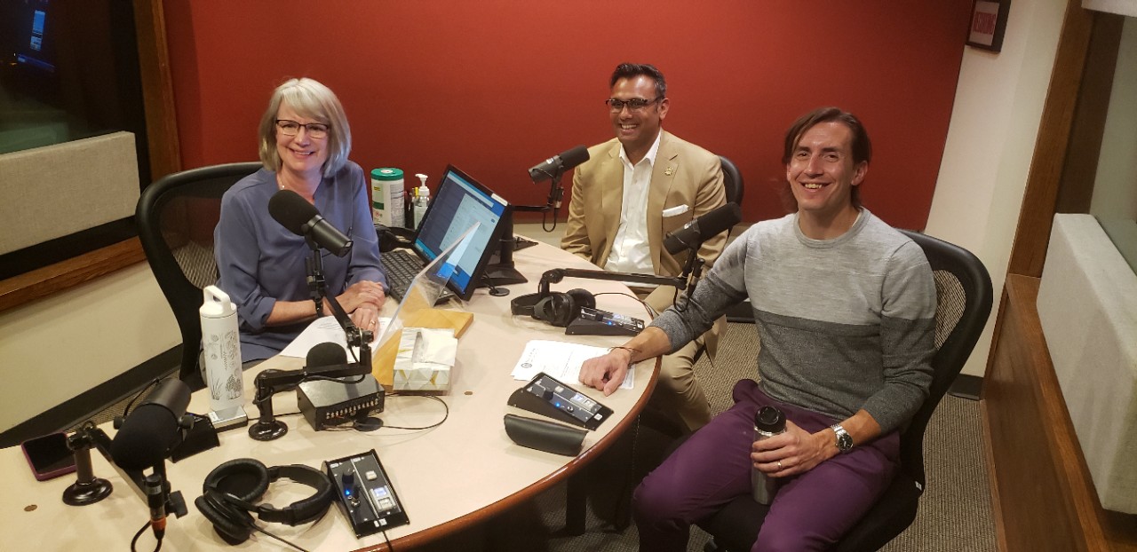 Lucy May behind a desk and Pavan Parikh and Nick Zingarelli join her on the outside side with microphones in front