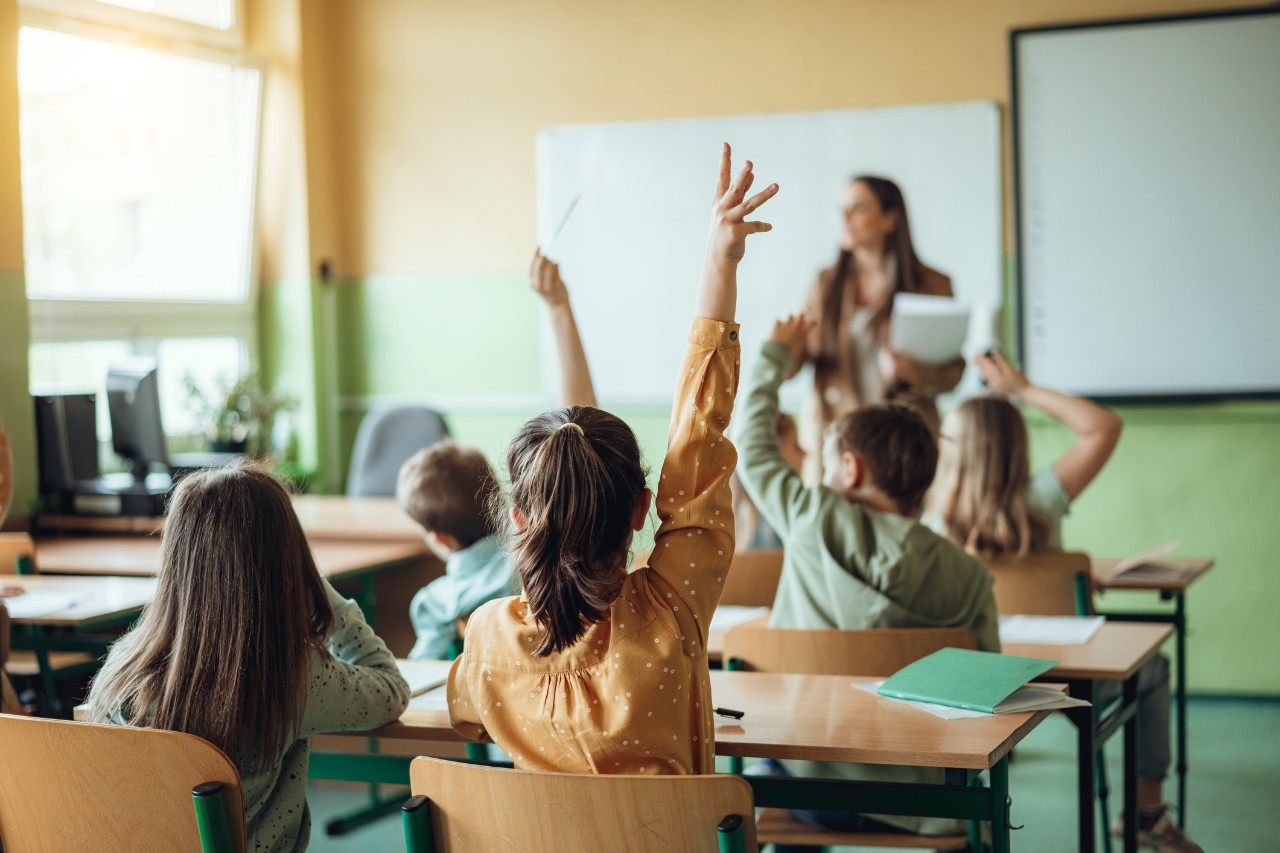 teacher at the front of the classroom and students with their hands raised