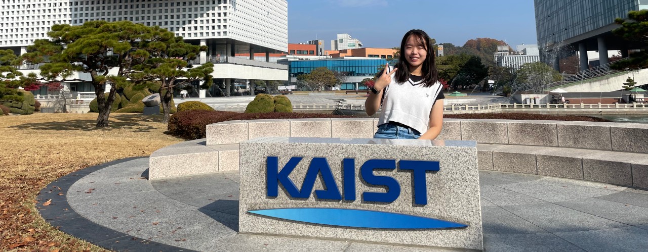 Woman stands behind KAIST sign and in front of several technical park buildings in South Korea.