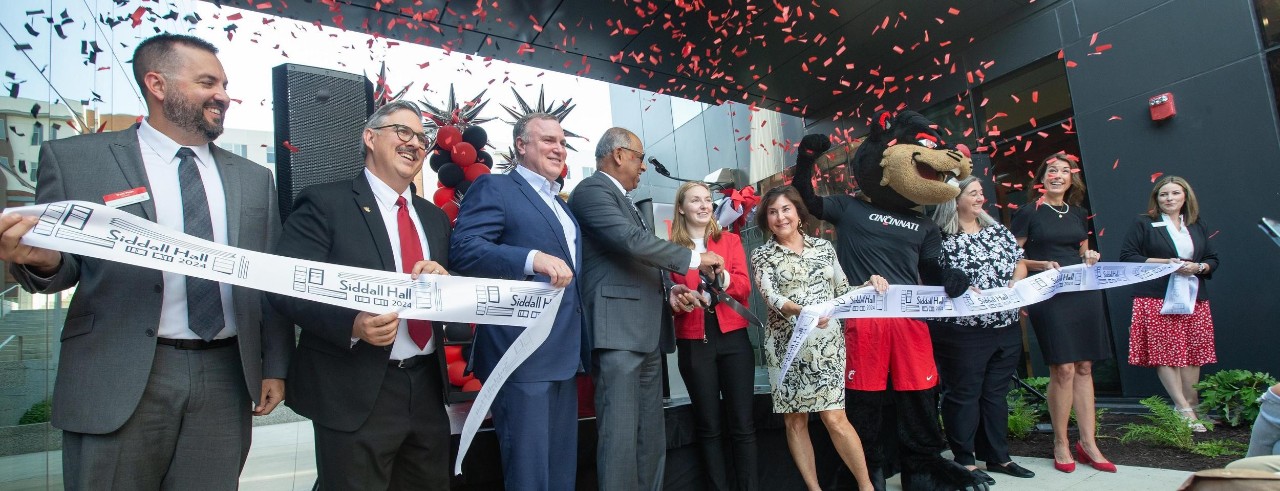 Ten UC administrators including President Pinto, the student body president Madison Wesley and the Bearcat stand and hold a ribbon for a ribbon cuting.  Confetti cannon goes off.