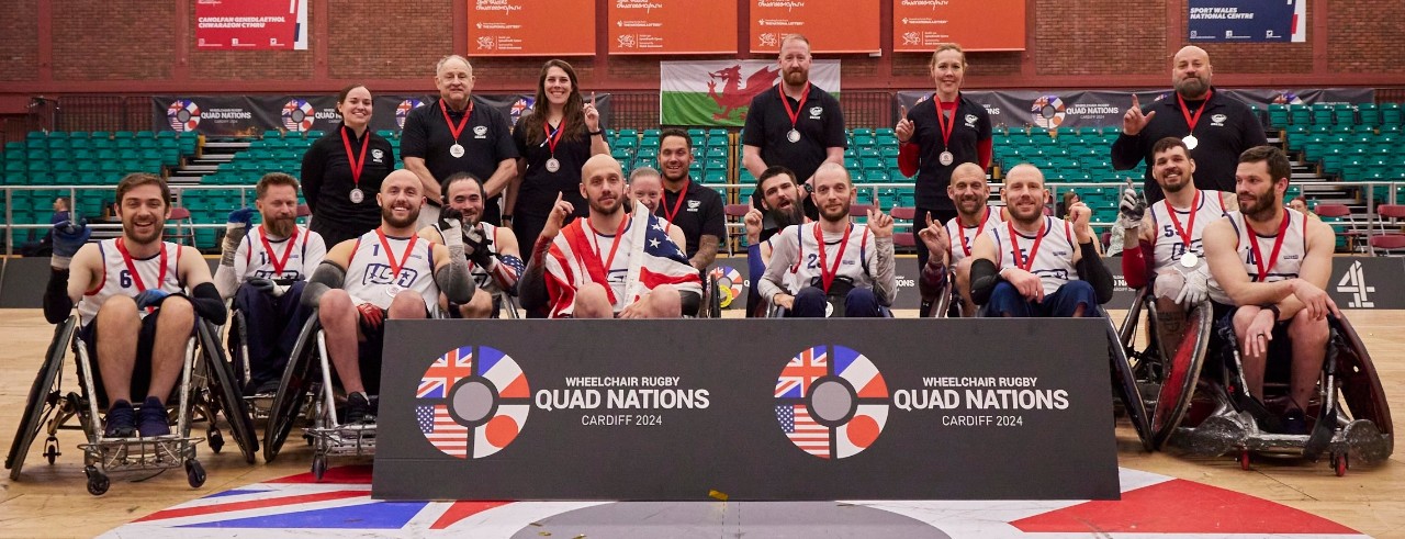 The Team USA wheelchair rugby team takes a team photo on a court following a tournament. Staff members stand behind players in wheelchairs, who all have medals around their necks.