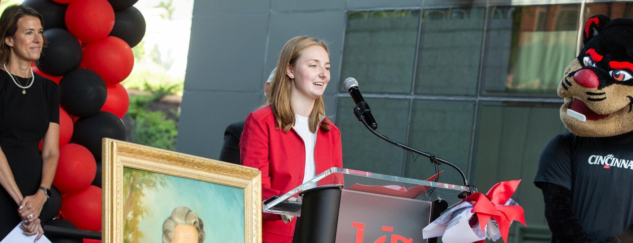 Dean Nicole Mayo stands to the left of a photo of Helen Siddall while Madison Wesley stands at the podium to speak.  The Bearcat Mascot stands nearby