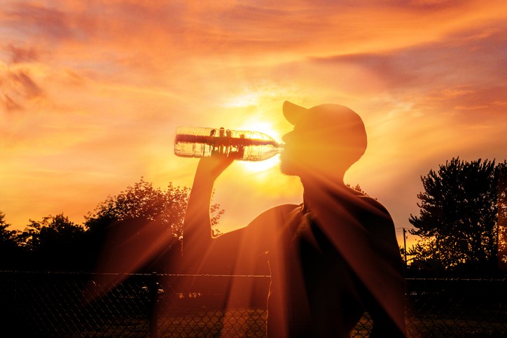 Silhouette of a man wearing a baseball cap drinking a bottle of water in front of trees and an orange sky and sun