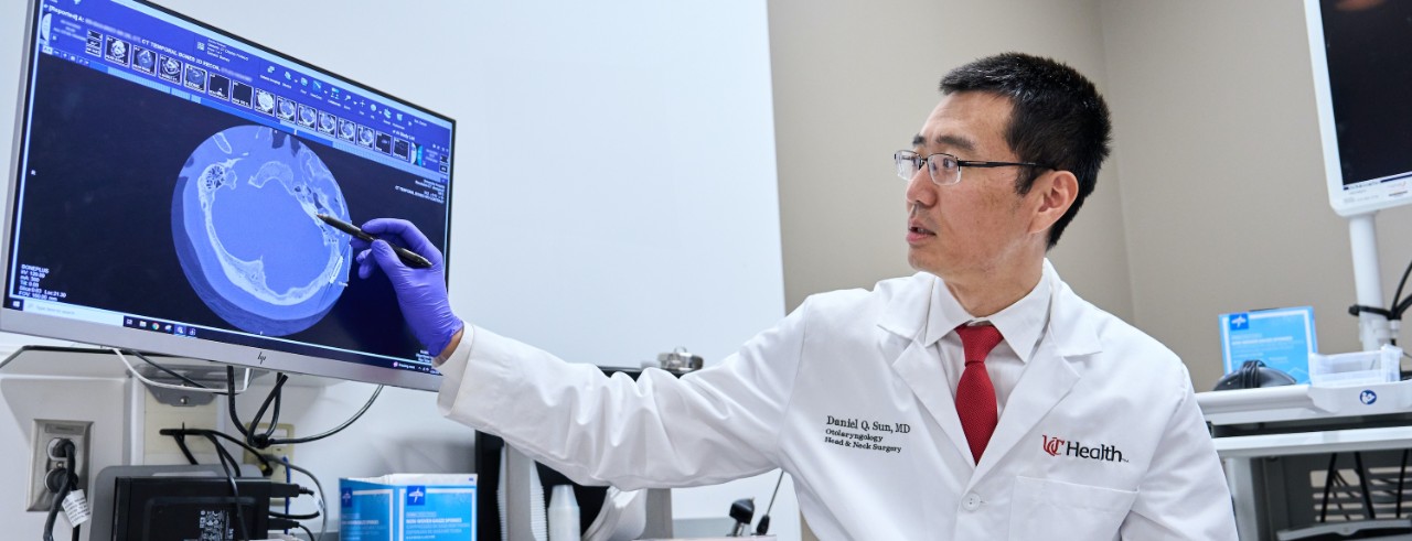 Wearing a white coat and glasses, Daniel Sun uses a pen to point at an area of a brain scan on a computer monitor