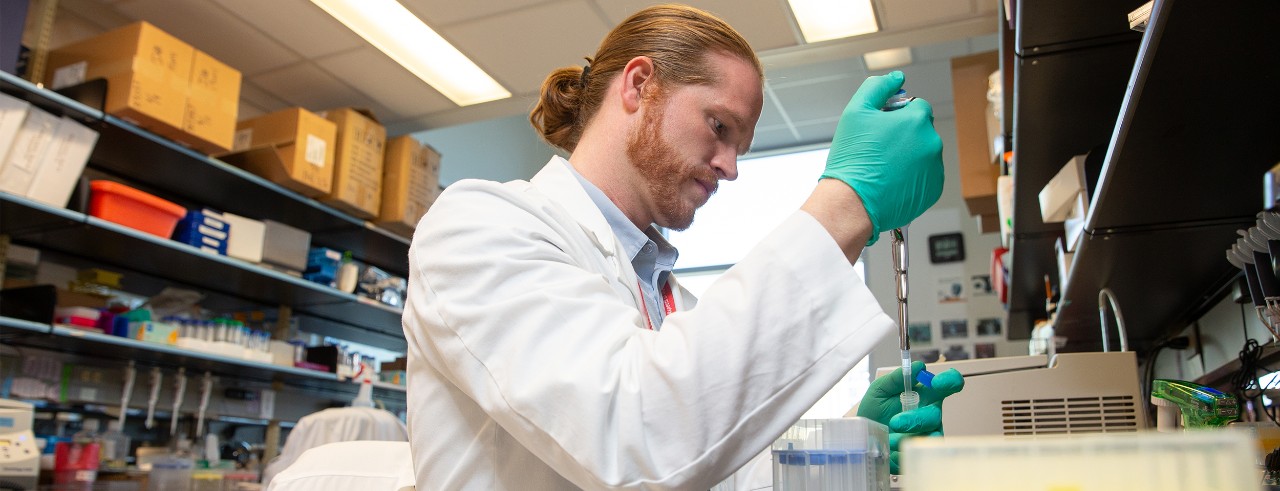 Eric Vick, wearing a white lab coat, pipettes a sample in a laboratory