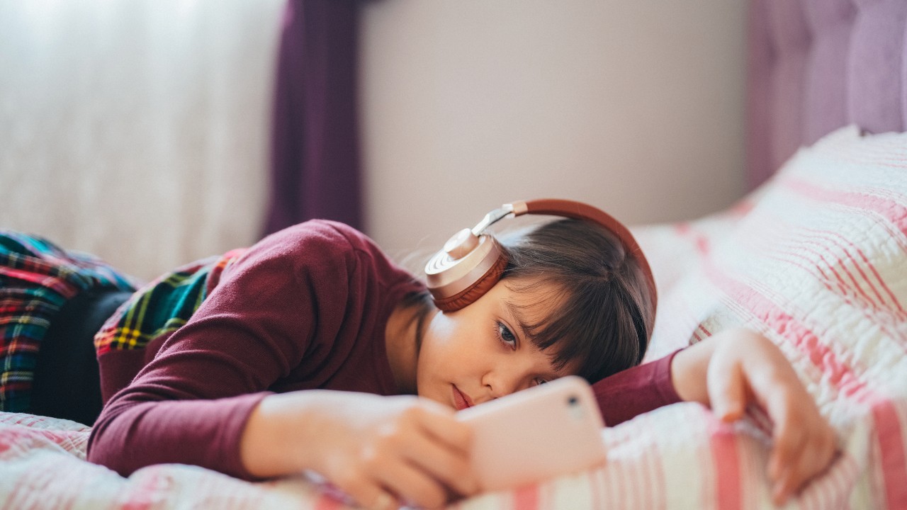 Gen Alpha male on the bed with headphones and a cell phone watching something