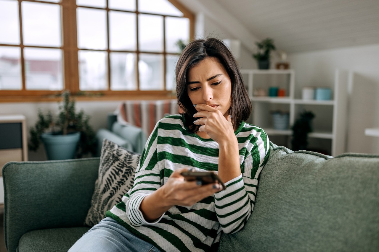 A woman with a concerned expression scrolls her phone on the couch.