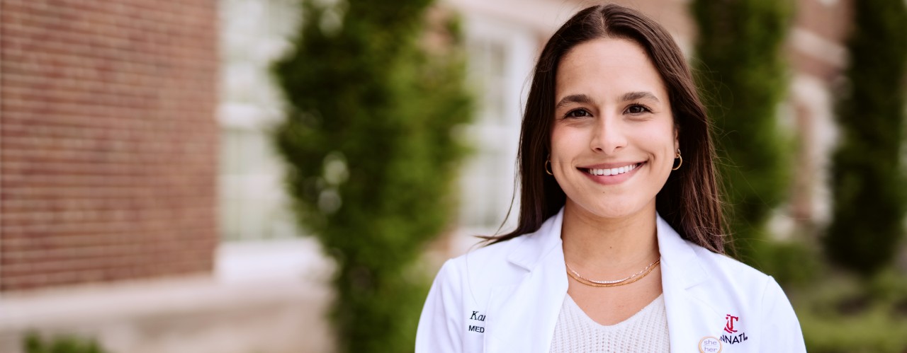 UC med student Kamala Nelson faces forward wearing a white medical jacket in front of a brick building and bushes.