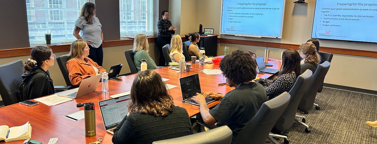 university faculty and staff sit around a large table looking at a screen with a presentation titled preparing for the proposal