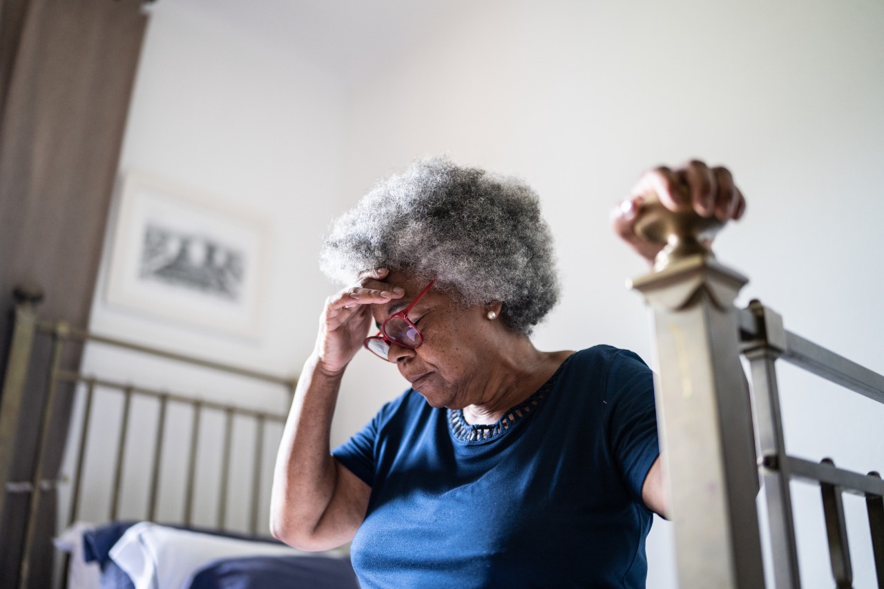 An elderly woman sitting on her bed holds her head because she has a headache