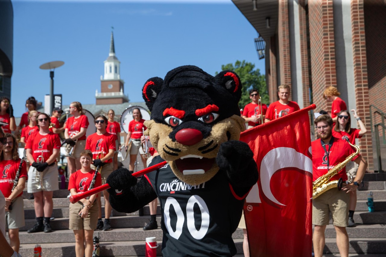 Bearcat Mascot holds UC flag and many members of the marching band play in the background