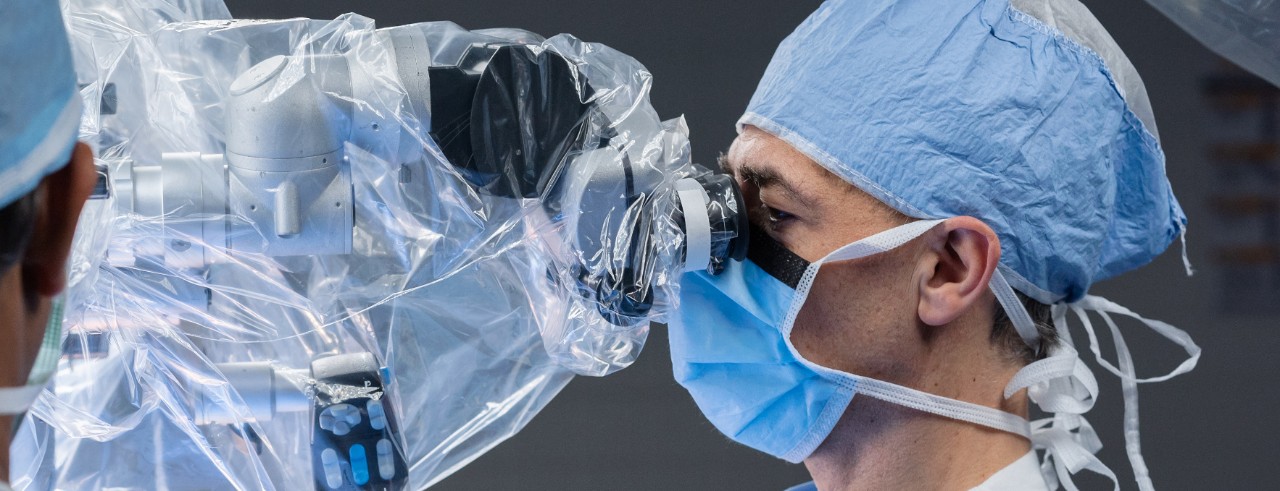 Norberto Andaluz wears a scrub cap and mask while looking through a microscope during surgery