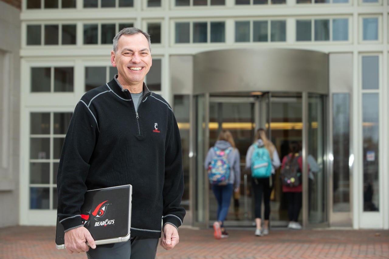 older man with laptop wearing UC swag standing in front of the College of Education on the UC campus.  Two students in the background walking into building