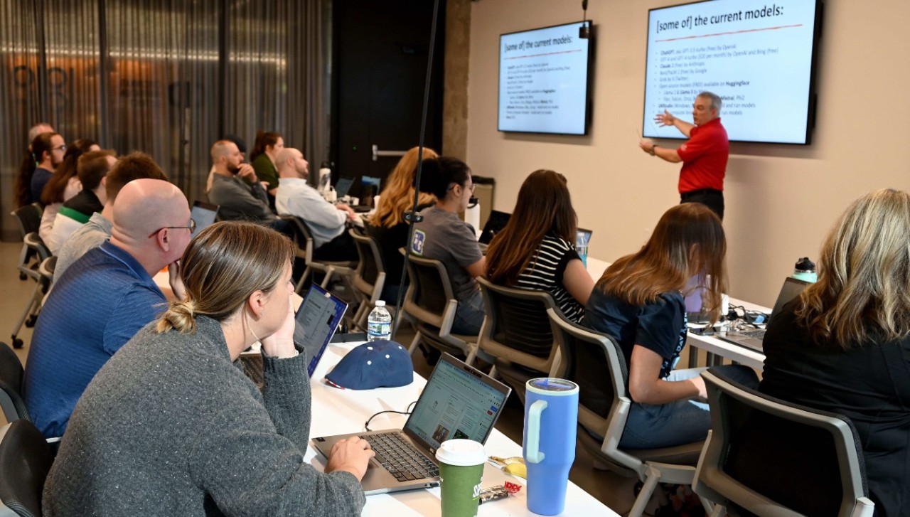 A man in a red shirt and black pants instructs a class full of professionals with laptops open.