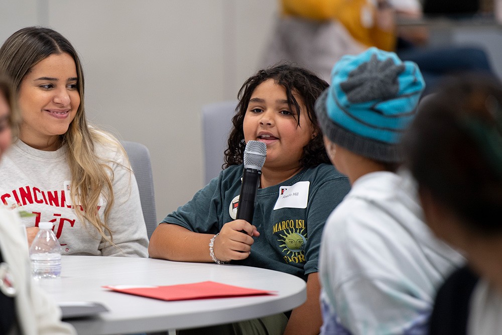 A child speaks into a microphone while other people look on.