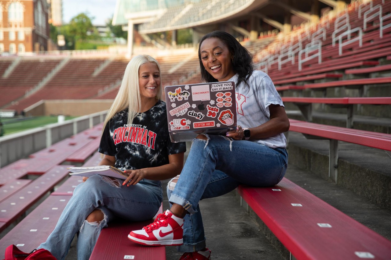 Two women students smiling in Nippert Stadium stands, using laptops and enjoying college life and the campus experience