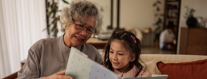 A grandmother sits on a couch with her granddaughter and reads her a book