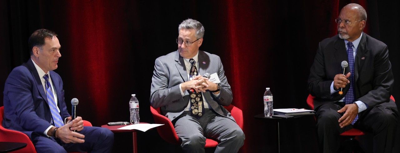 Three men in suits sit in chairs on a stage as part of a panel discussion.