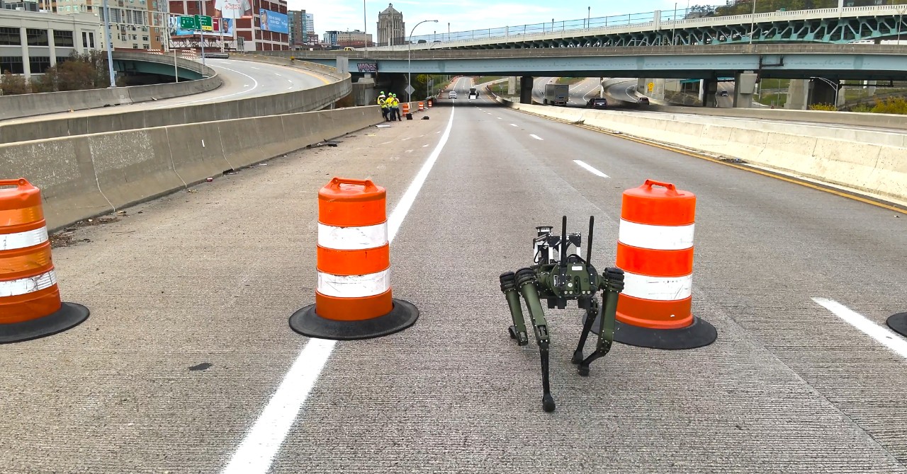 A robotic dog walks past orange barrels on a closed bridge.