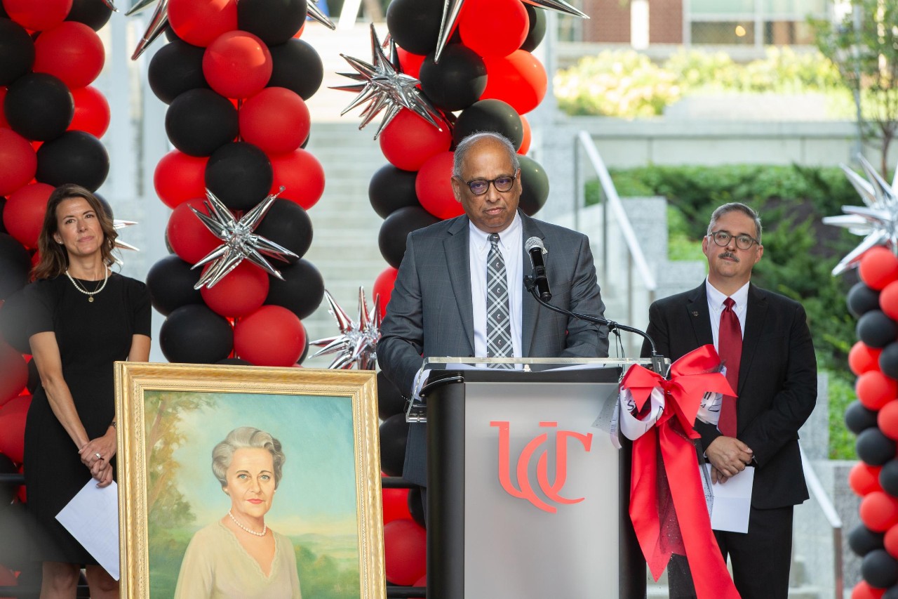 Neville Pinto stands at the podium while Bearcat Mascot stands nearby. Two UC administrators stand on either side of Pinto with the Bearcat Mascot standing afar