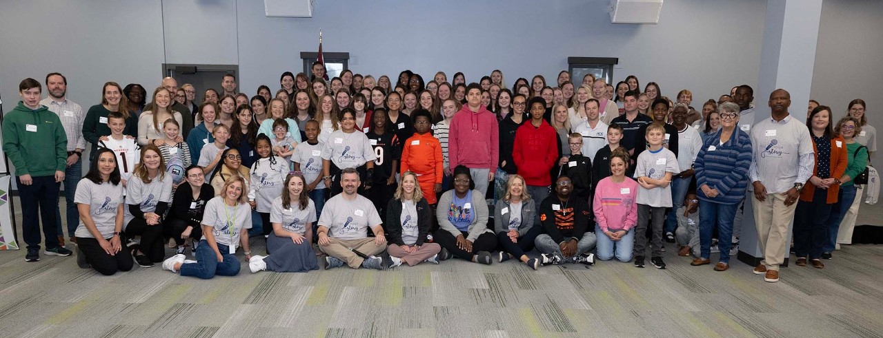 A group of children, teens and adults pose for a photo with some sitting or kneeling and others standing behind them.